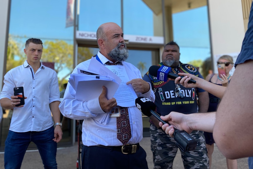 A man with a beard and wearing a tie holds a bundle of papers and speaks into two microphones. 