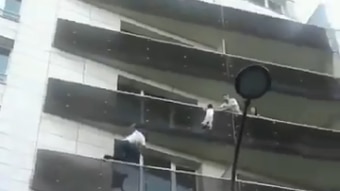 A man is scaling a residential apartment building to save a young boy who is hanging from a balcony on the fourth storey.