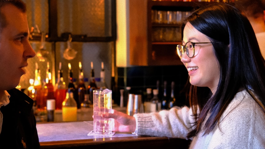 A young woman is sitting in a wine bar holding a glass of soda water as she chats with a young man.  