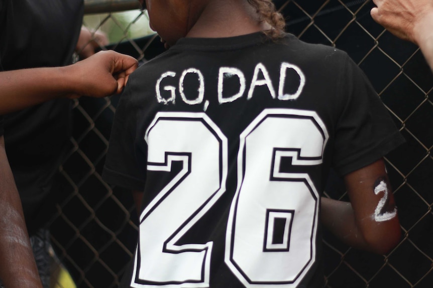 Child stands in front of a fence with a Magpies t-shirt saying 'go dad'.