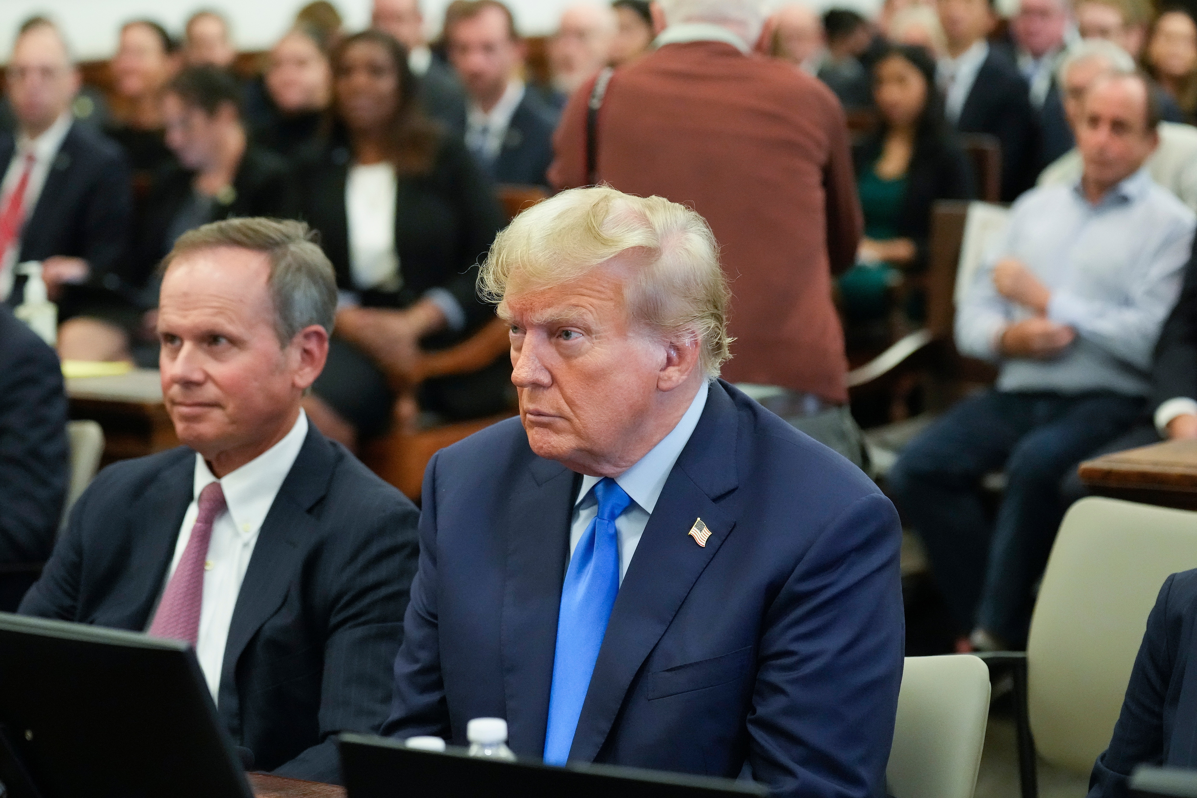 Two men in suits and ties sit next to each other in a courtroom, in front of a large group of other people.