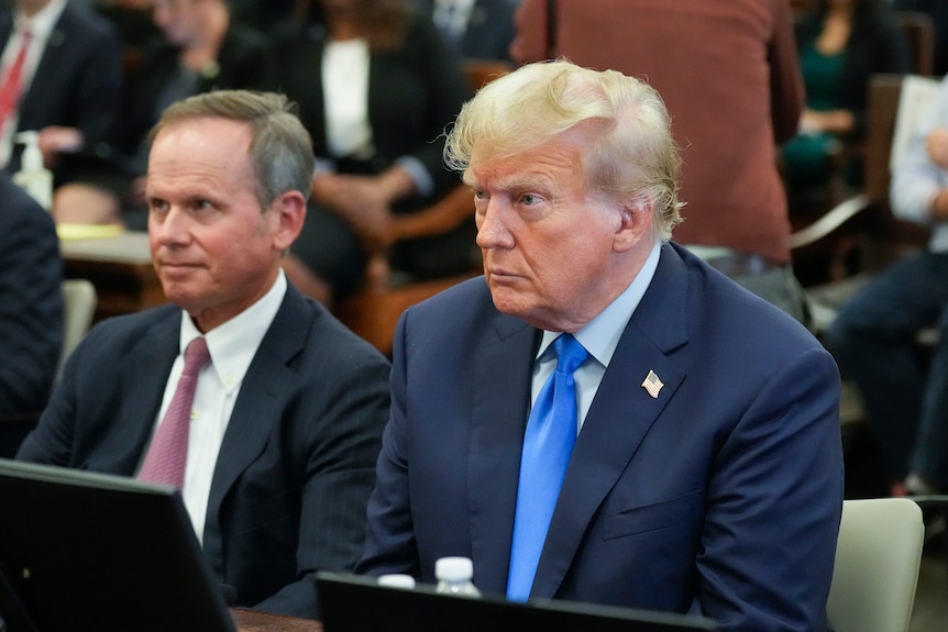 Two men in suits and ties sit next to each other in a courtroom, in front of a large group of other people.