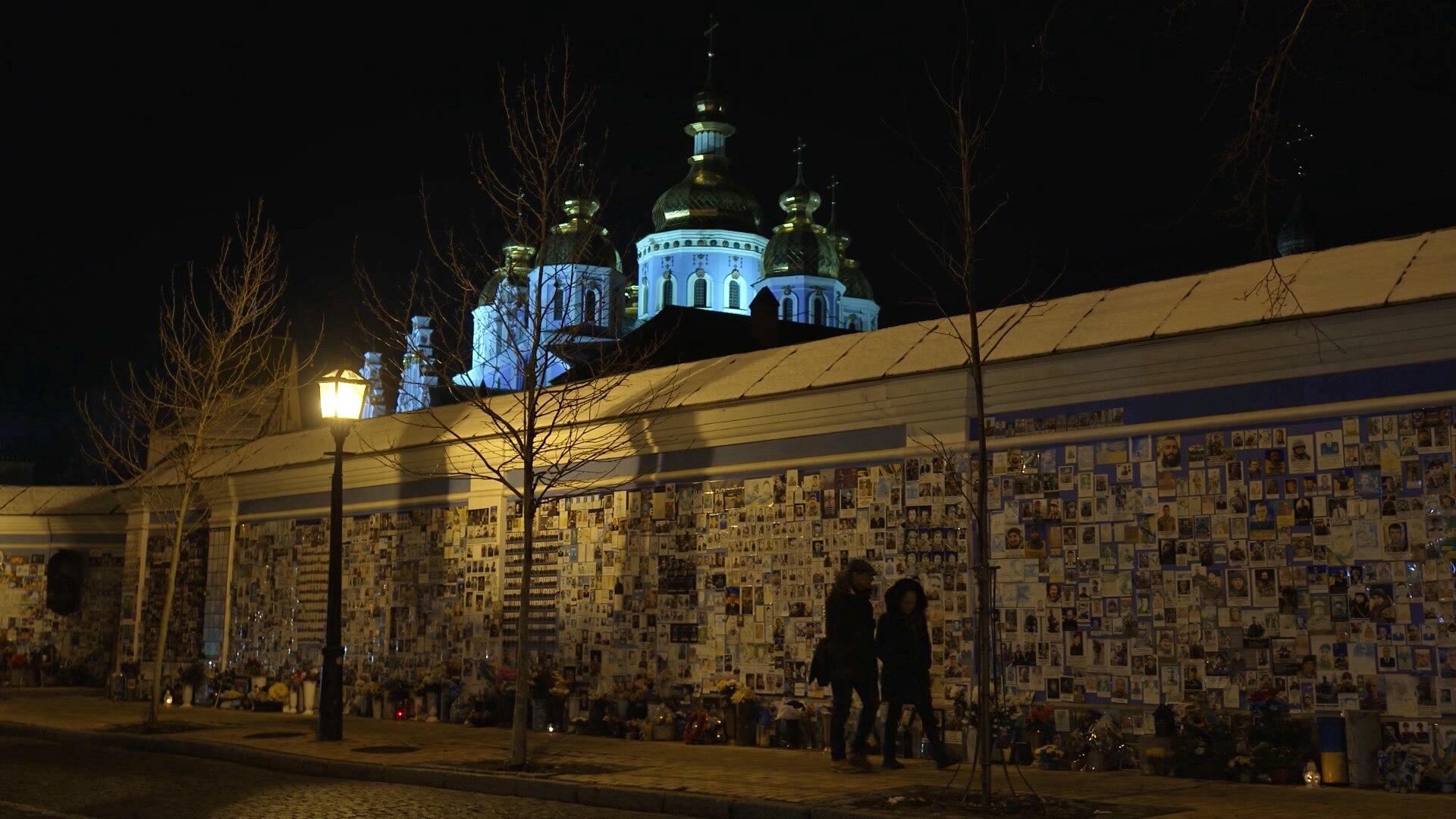 A high wall covered in photos of people. Two people walk by it at night. A large building behind the wall is illuminated.