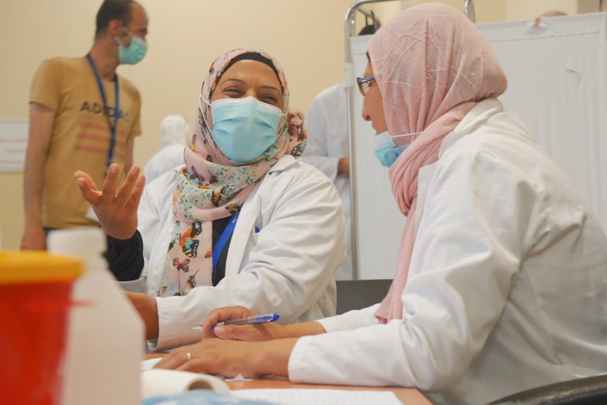 Two women wearing masks, hijabs and medical coats talk as a man behind them lines up for vaccination.