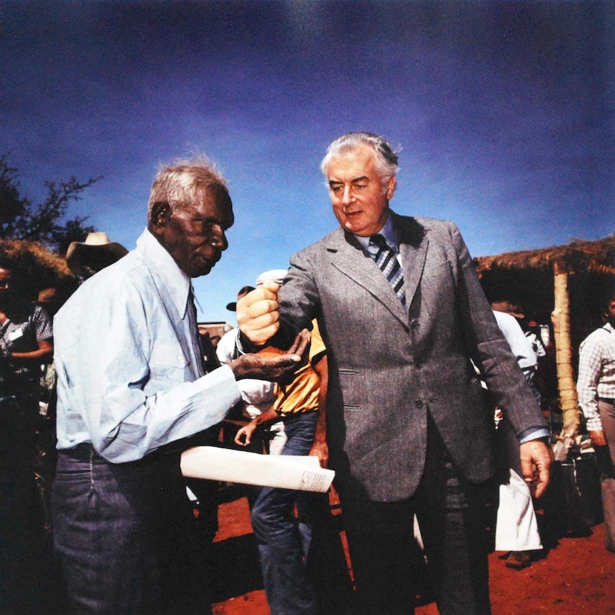 Gough Whitlam in grey suit, blue striped tie, pours soil into Indigenous man Vincent Lingiari who wears blue shirt, tie. 