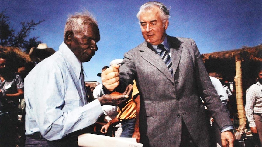 Gough Whitlam in grey suit, blue striped tie, pours soil into Indigenous man Vincent Lingiari who wears blue shirt, tie. 