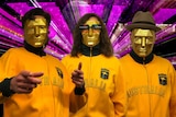 Three men appear in yellow jackets with Australia written across them. They are each wearing gold masks.