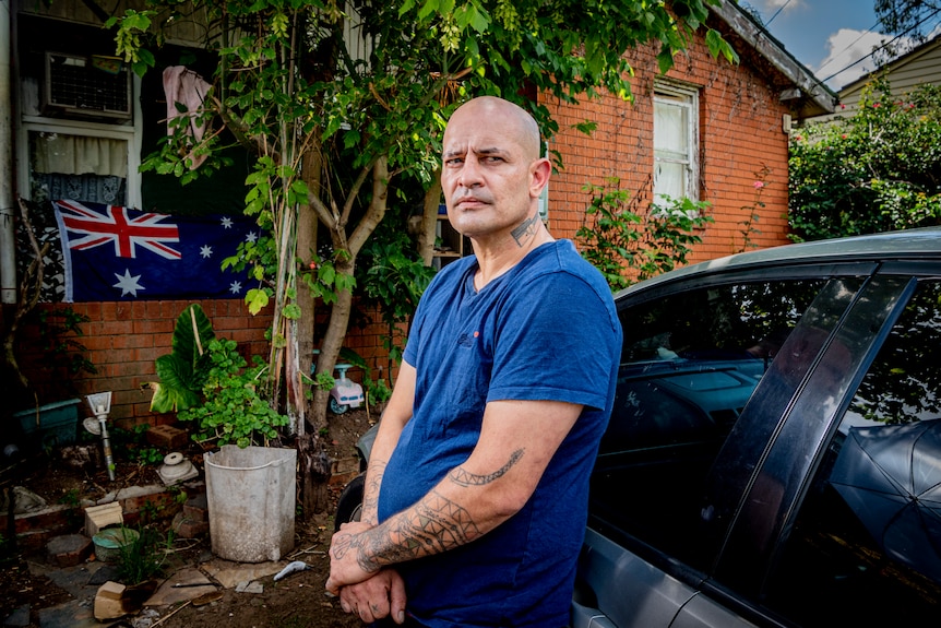 Man in a blue shirt leans against a car outside a red brick home.