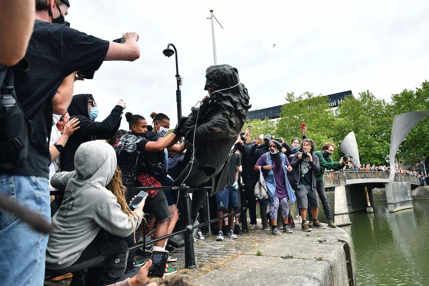 a crowd is packed behind a small fence, pushing a large statue of a 17th century man over it and into a river on the other side.