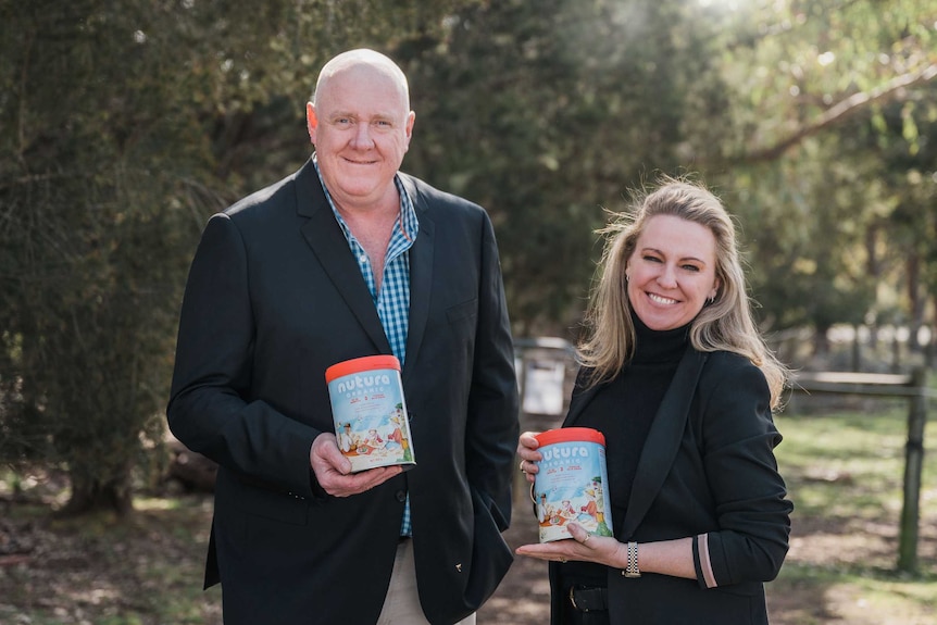 A man and a woman hold two cans of infant formula in a garden