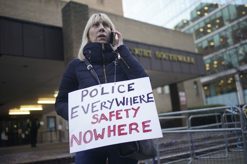 A blonde woman speaks on the phone. she stands outside a building holding a sign that reads "police everywhere, safety nowhere"