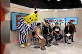 A television set with four seated people talking. Behind them are six dressed up characters, including the Bananas in Pyjamas.