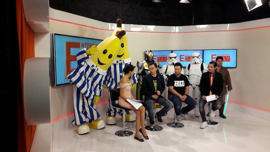 A television set with four seated people talking. Behind them are six dressed up characters, including the Bananas in Pyjamas.