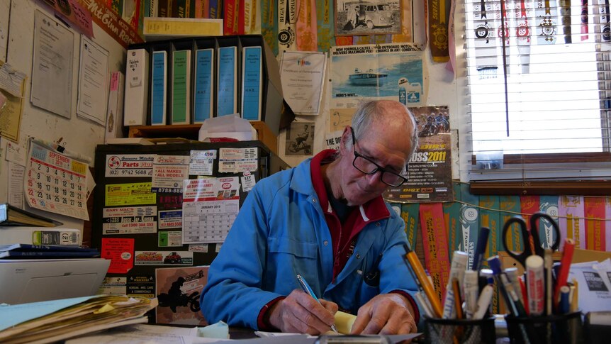 owner Wayne Flynn sits at a work desk littered with stationary. His office is filled with paper, magnets and ribbons.