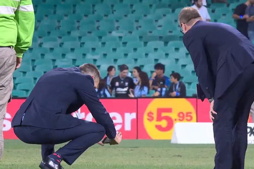 A man in a suit bends down and photographs a patch of grass, with two other men watching, empty seats in the background.