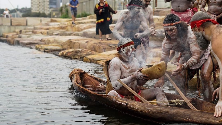 An Indigenous man in a canoe hands a bowl of fire to other men on the shore