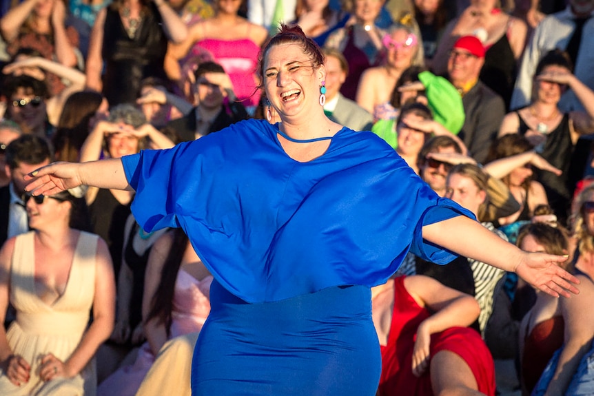 A smiling woman in a blue dress smiles in front of a crowd of people.