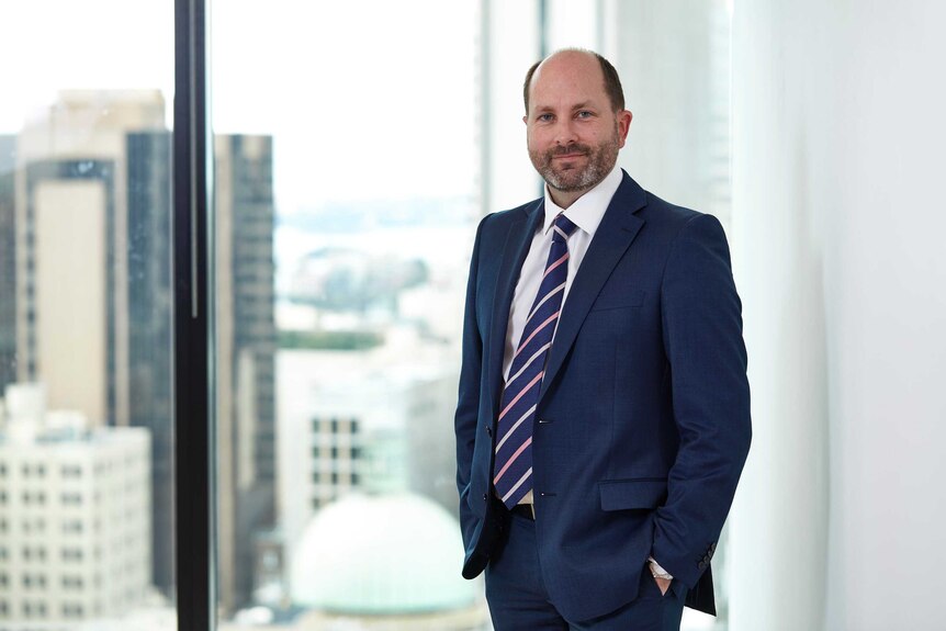 A man in a suit and a blue and pink striped tie standing in front of a window with views of city skyline