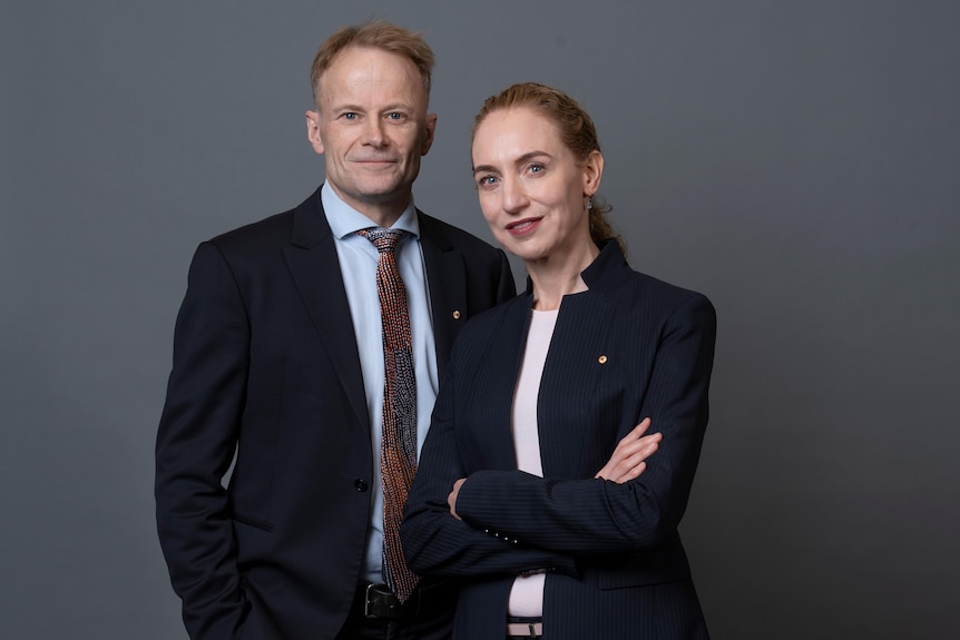 A man and woman stand side-by-side wearing suits. 