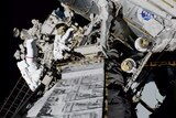 Two astronauts in white spacesuits cling to the side of a spacecraft with a NASA logo on the side.