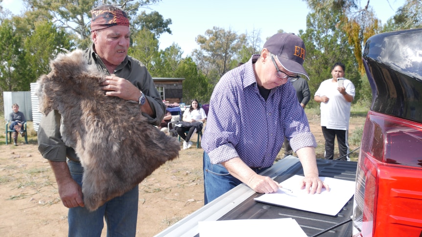 Indigenous elder wearing a kangaroo skin watches as a non-indigenous woman signs legal documents on the back of a ute. 