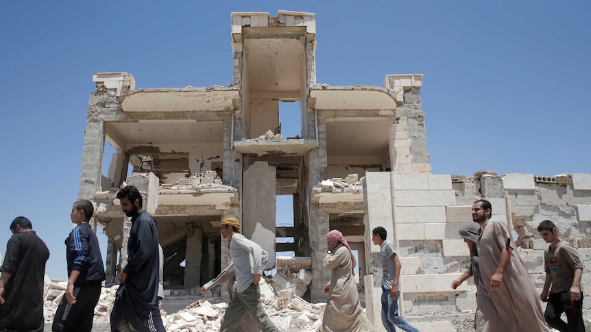 Islamic State prisoners dressed in western clothes or traditional robes walk past a ruined building.