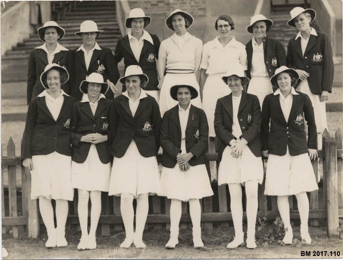 13 mulheres em uniformes de críquete (vestidos brancos na altura dos joelhos com blazers) posam para uma fotografia. 