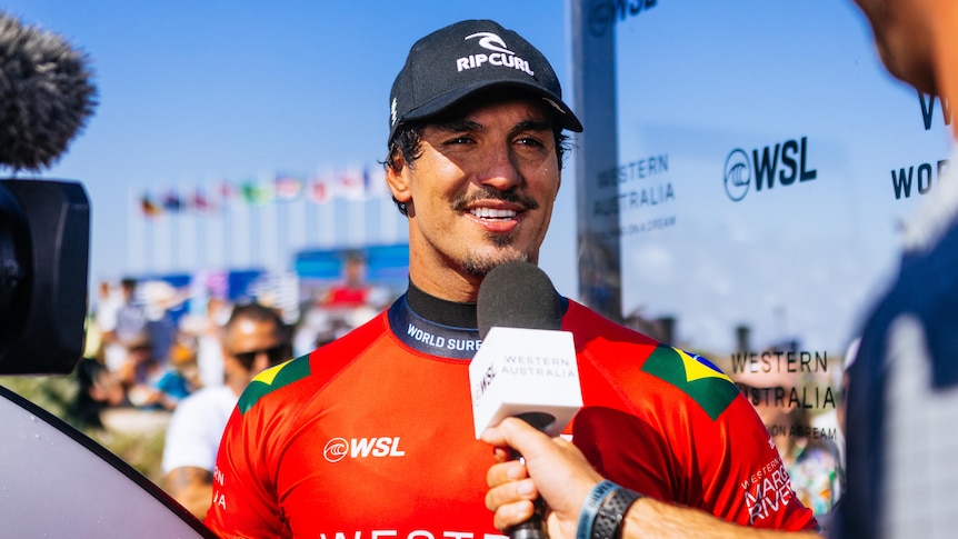 A man in an orange rashie and black cap being interviewed on camera