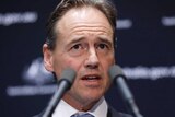 Greg Hunt gestures at a lecturn with two microphones in the foreground at a press conference