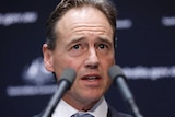 Greg Hunt gestures at a lecturn with two microphones in the foreground at a press conference