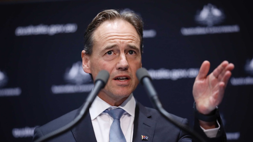 Greg Hunt gestures at a lectern with two microphones in the foreground at a press conference