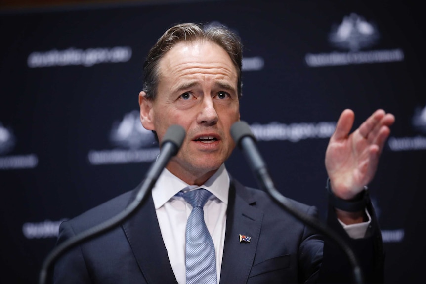 Greg Hunt gestures at a lecturn with two microphones in the foreground at a press conference