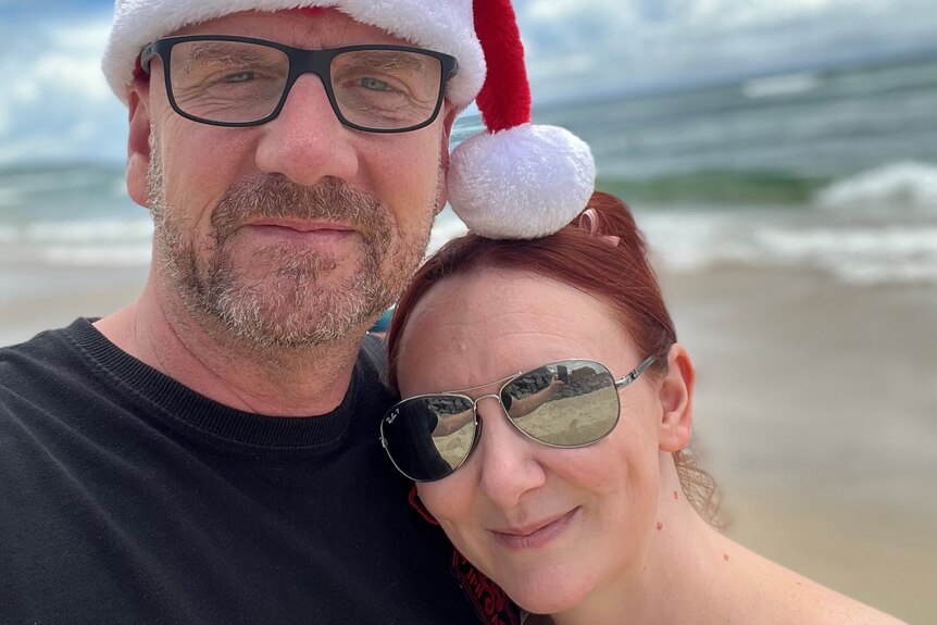 A man and a woman stand together on a beach, smiling. The man has a Christmas hat on.