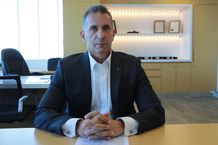 Paul Papalia wears a suit and sits at his desk with his hands clasped