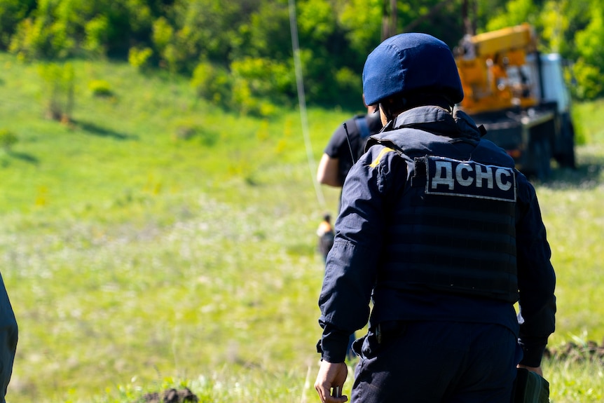 A man in a helmet and navy safety suit walks through a field 