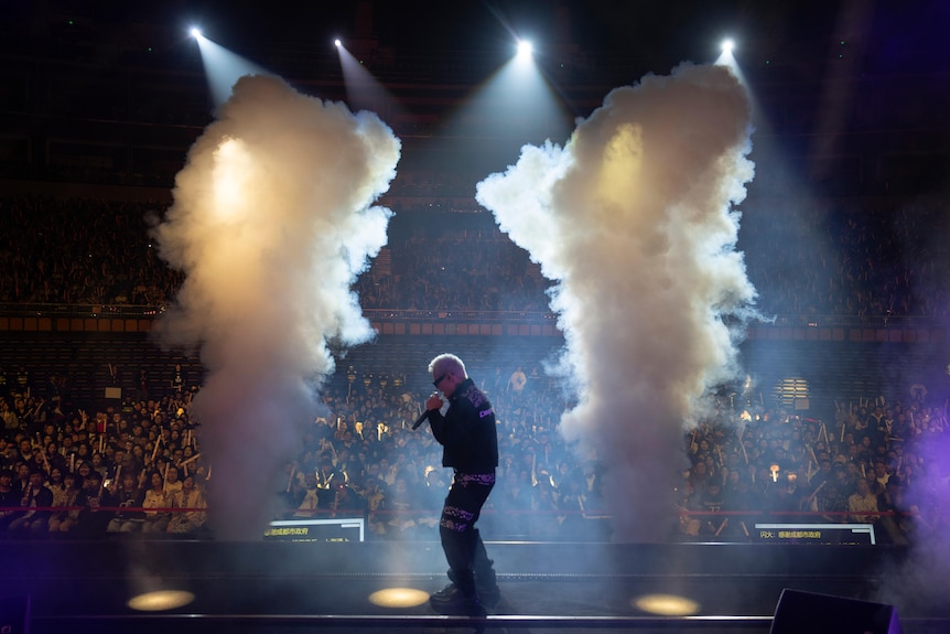 舞台上，一名金发碧眼的年轻中国男子对着麦克风唱歌，烟雾从他周围的两根柱子中升起。