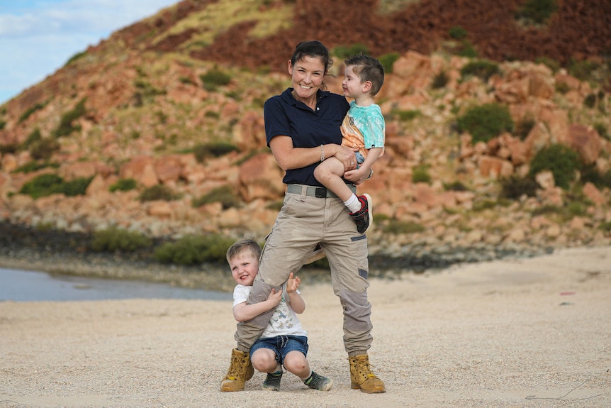 Smiling woman in black T-shirt with company logo, khaki pants, boots, boy clings to her leg, holds another, beach, red rocks.