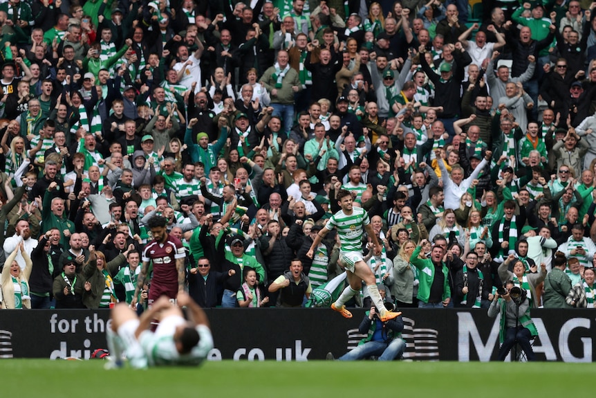 Football players in green and white jump up and down in celebration on a pitch as a packed crowd behind them cheers and celebrates