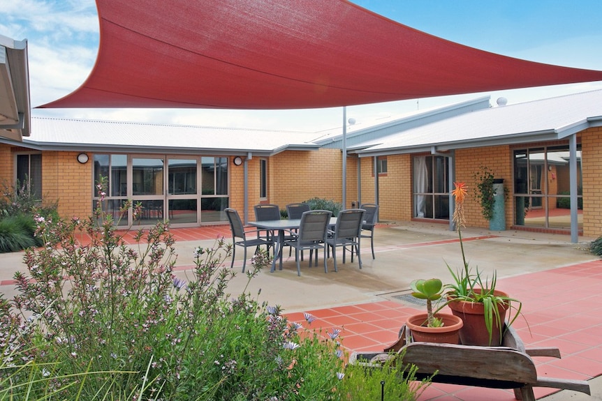 A court yard at an aged care home with a red shade protector and a glass table surrounded by chairs.