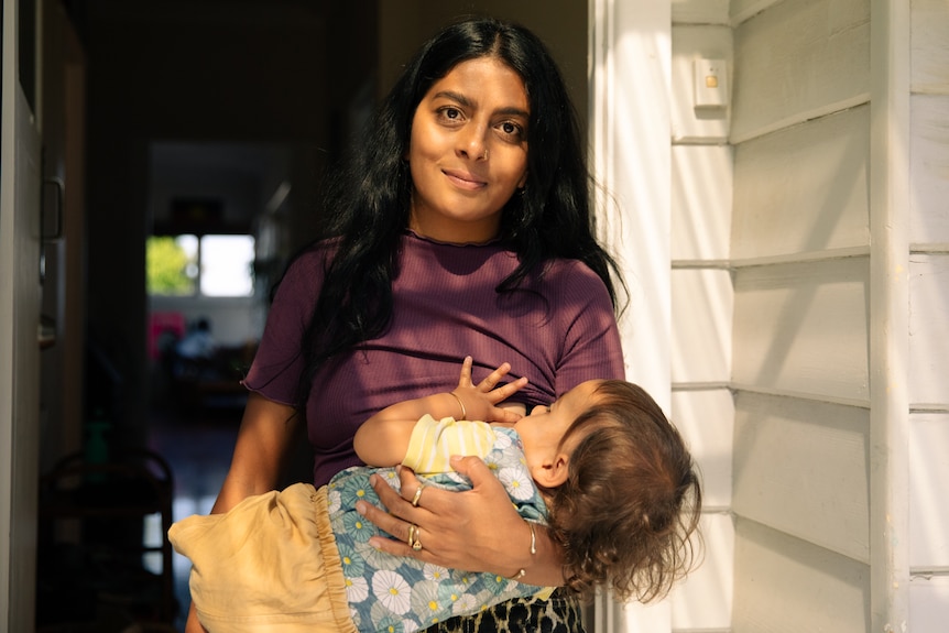 A woman standing at her front door, breastfeeding her baby.