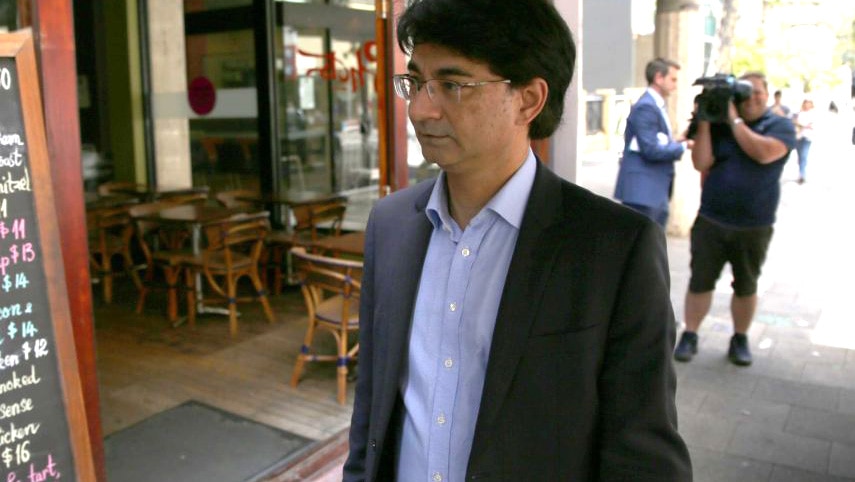 Lloyd Rayney in a blue suit walks in front of a cafe as media crews film him in the background.