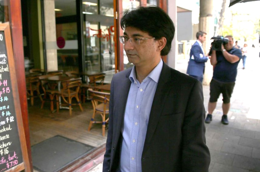 Lloyd Rayney in a blue suit walks in front of a cafe as media crews film him in the background.