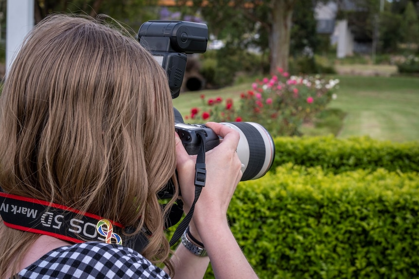 A photograph taken over the shoulder of a woman with long brown hair taking a photograph in a garden.
