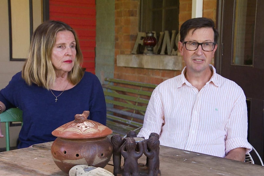 A woman sits by a picnic table on a porch with her husband discussing a charity.