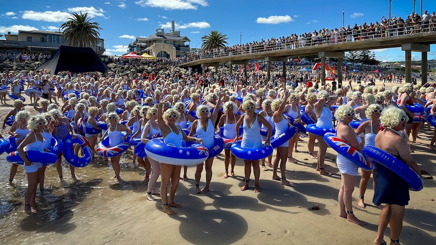 Hundreds of people wearing white swimwear, blonde curly wigs and blue swim rings stand along the sand and jetty