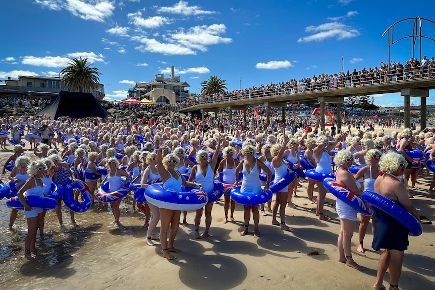 Hundreds of people wearing white swimwear, blonde curly wigs and blue swim rings stand along the sand and jetty