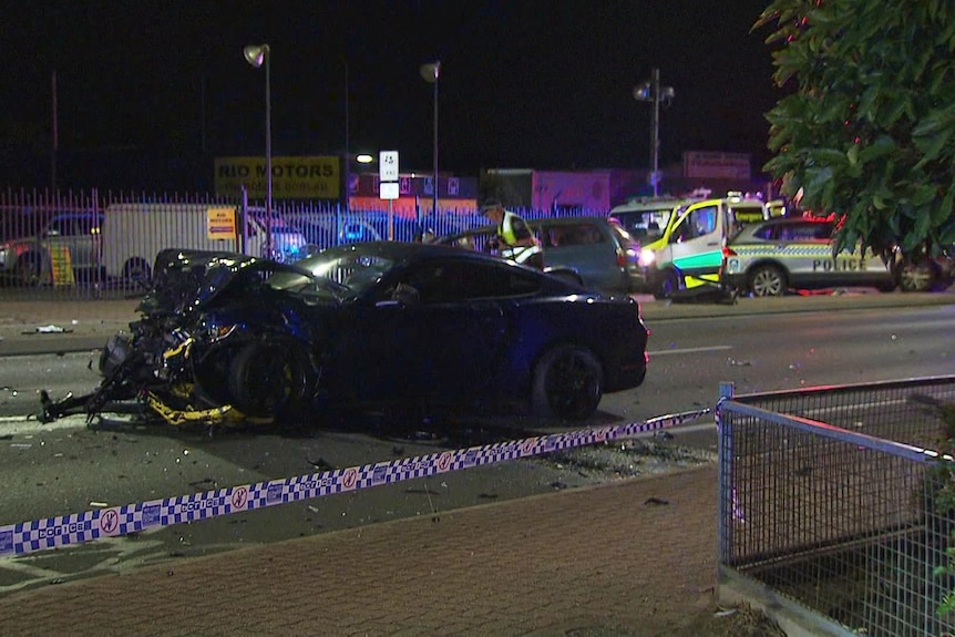 A severely damaged black car on a road. Police tape is in the foreground, and ambulances are in the background