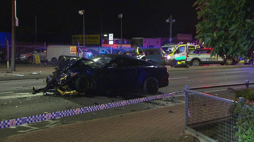 A severely damaged black car on a road. Police tape is in the foreground, and ambulances are in the background