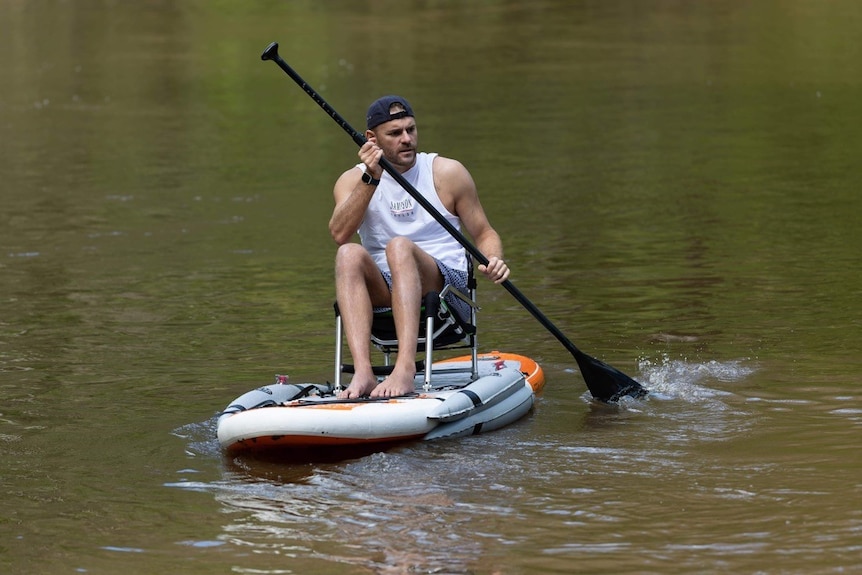 A man in white singlet sitting and paddling an inflatable paddle board on flat waters