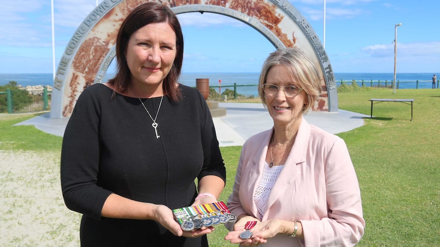 Two women stand in front of a war memorial at the beach holding their medals of service.